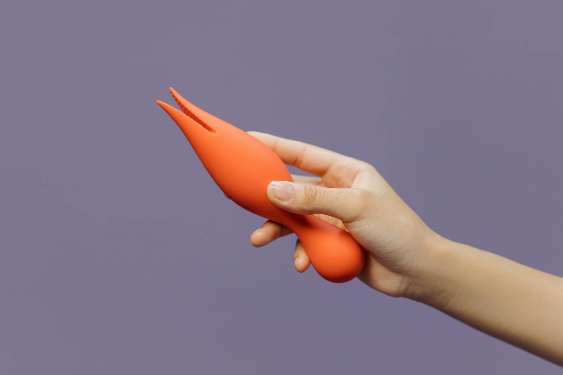 Eine Hand hält einen orangen Lemon Vibrator gegen einen minimalistischen lila Hintergrund.