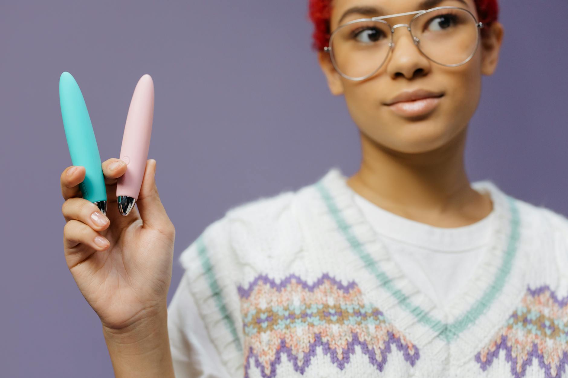 Junge Frau mit Brille hält zwei farbenfrohe Vibratoren vor einem violetten Hintergrund